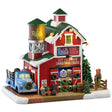 This image shows what this Lemax Harvest Crossing lighted building looks like on a white background. It was designed to look like a red barn with a green roof covered in snow. There's a water tower that says "Reid's" and a sign on the side of the building that says "Reid's Trees & Wreaths. Cut Your Own Christmas Trees." Inside the building, there are rows of decorated wreaths. There's a blue truck on the side of the building and a lot full of trees on the other side.