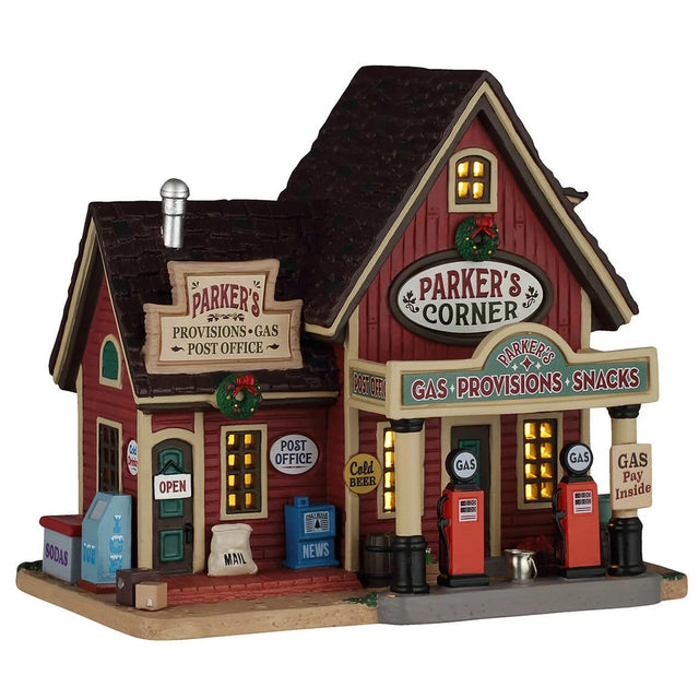 This image shows what this Lemax building looks like on a white background. It was designed to look like a red cottage turned into a gas station / post office. There are signs that read "Parker's Provisions, Gas, Post Office," "Parker's Corner," "Parker's Gas, Provisions, Snacks," "Cold Beer" and "Gas - Pay Inside!" There are gas pumps, freezers and a newspaper vending machine.