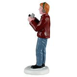 This image shows one of the figurines. This one is Dad holding a camera. He has blonde hair and is wearing green earmuffs. He's wearing a maroon colored jacket, blue jeans and black shoes.