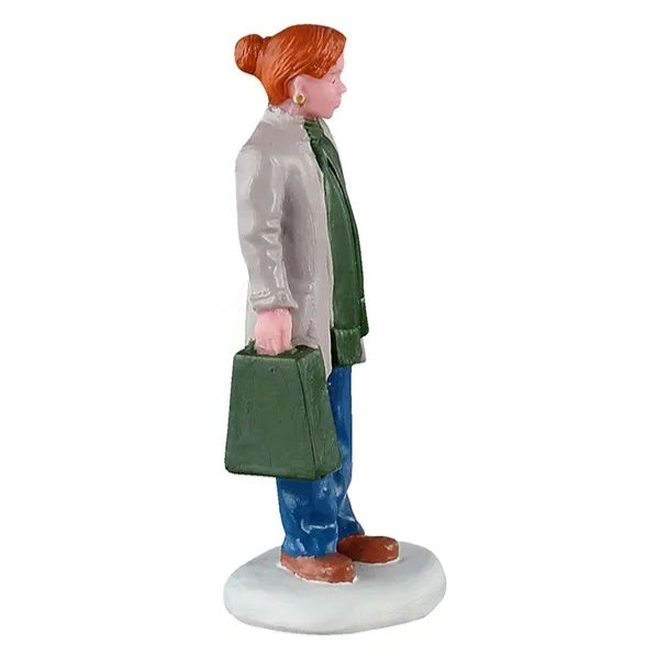 This image shows one of the figurines. This one is Mom holding a green shopping bag. She has orange hair and is wearing gold earrings, a gray jacket, a green scarf, blue jeans and brown shoes.