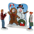 This image shows what these Lemax figurines look like on a white background. The set of three is designed to look like two kids are posing behind a face-in-hole board while Dad takes pictures. Mom is watching and holding a shopping bag.