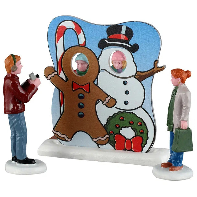 This image shows what these Lemax figurines look like on a white background. The set of three is designed to look like two kids are posing behind a face-in-hole board while Dad takes pictures. Mom is watching and holding a shopping bag.
