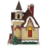 This image shows what this building looks like from the side. There's a stained glass window and a tree that's decorated with ornaments. The roof and the ground are covered in snow.