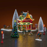 This is a lifestyle image showing what this Lemax house set looks like on a fake background. The set was designed to look like a Christmas-themed barn turned into a Christmas store that sells garlands, trees and wreaths. There are snow covered trees and a lighted tree. Santa's sleigh is being led by reindeer while an elf loads presents into the sleigh. There are elves all over, as well as lampposts.