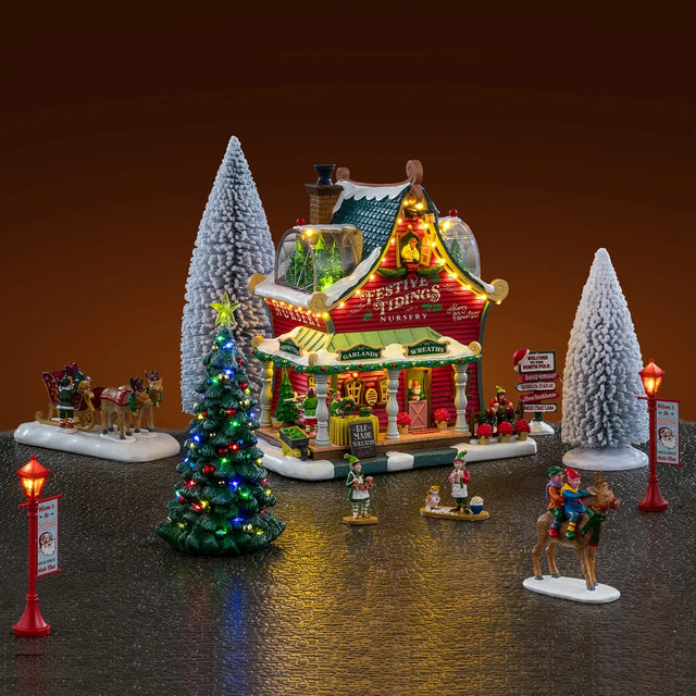 This is a lifestyle image showing what this Lemax house set looks like on a fake background. The set was designed to look like a Christmas-themed barn turned into a Christmas store that sells garlands, trees and wreaths. There are snow covered trees and a lighted tree. Santa's sleigh is being led by reindeer while an elf loads presents into the sleigh. There are elves all over, as well as lampposts.