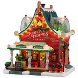 This image shows what this Lemax Santa's Wonderland lighted building looks like on a white background. It was designed to look like a red house turned into the Festive Tidings Nursery. On the porch, there's a table with elf made wreaths for sale. On the side of the house, there's an elf decorating wreaths. There are two dome windows on the roof where you can see some trees inside.