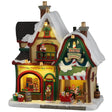 This image shows what this Lemax building looks like on a white background. It was designed to look like a Christmas-themed store. The roof and sidewalks are covered in snow. There are signs that read "North Pole Christmas Supply," "Ribbons, Bows, Wrapping Paper, Holly, Ornaments" and "2 for 1 Ornaments." There are elves all over working and loading things into a sleigh being pulled by a reindeer.