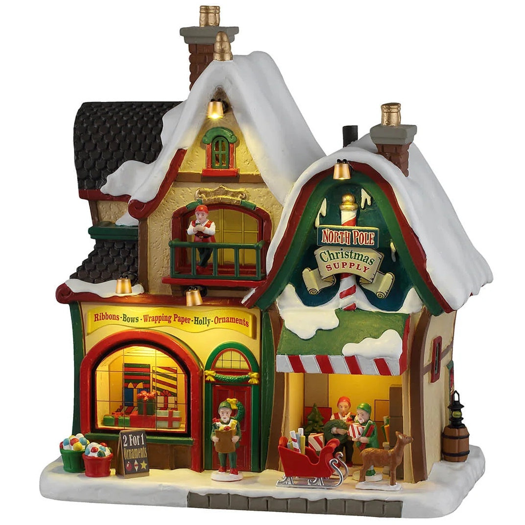 This image shows what this Lemax building looks like on a white background. It was designed to look like a Christmas-themed store. The roof and sidewalks are covered in snow. There are signs that read "North Pole Christmas Supply," "Ribbons, Bows, Wrapping Paper, Holly, Ornaments" and "2 for 1 Ornaments." There are elves all over working and loading things into a sleigh being pulled by a reindeer.
