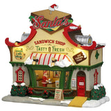 This image shows what this Lemax building looks like on a white background. It was designed to look like a restaurant. On the roof, there's a large, decorative, fake sandwich with a pick in it. The pick has a Santa's hat on it. There's a large window in the front where you can see the inside. Santa is picking up his food from the counter inside. There's snow on the roof and on the sidewalk.
