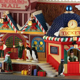This is an up close view of the lighted building. There are presents and suitcases on the sidewalk and lots of elves working.