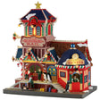 This image shows what this Lemax Santa's Wonderland lighted building looks like on a white background. It was designed to look like a multi-colored train station decorated for Christmas. There are candy cane and red bow accents everywhere. There are two penguins standing near the front door and one penguin is buying tickets from the ticket booth.