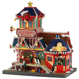 This image shows what this Lemax Santa's Wonderland lighted building looks like on a white background. It was designed to look like a multi-colored train station decorated for Christmas. There are candy cane and red bow accents everywhere. There are two penguins standing near the front door and one penguin is buying tickets from the ticket booth.