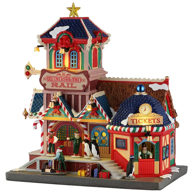 This image shows what this Lemax Santa's Wonderland lighted building looks like on a white background. It was designed to look like a multi-colored train station decorated for Christmas. There are candy cane and red bow accents everywhere. There are two penguins standing near the front door and one penguin is buying tickets from the ticket booth.
