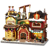 This image shows what this Lemax building looks like on a white background. It was designed to look like one building that has been split into two shops: The Holly Jolly Chocolatier and Mrs. Claus' Coffee Shop. There are windows at the chocolate shop and you can look in and see all the different candies on display. The whole building is decorated with wreaths and garlands and candy cane accents. At the coffee shop, there's a woman working behind the counter and some elves outside helping.