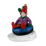 This figurine shows an elf in a blue and black inner tube in the snow. The elf is wearing a red hat with a gold bell on the end. He's also wearing a green scarf, a purple sweater, green gloves, orange pants and red shoes.