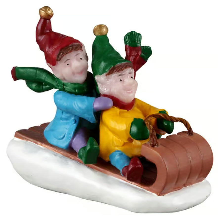 This image shows what this Lemax Santa's Wonderland figurine looks like on a white background. It was designed to look like two elves riding a brown toboggan downhill. The elf in front is wearing a yellow shirt, a green hat with a gold bell, green gloves, a red/green scarf, blue pants and brown shoes. The elf in back is wearing a blue jacket, a red hat with a gold bell, purple gloves, a green scarf, purple pants and green shoes.
