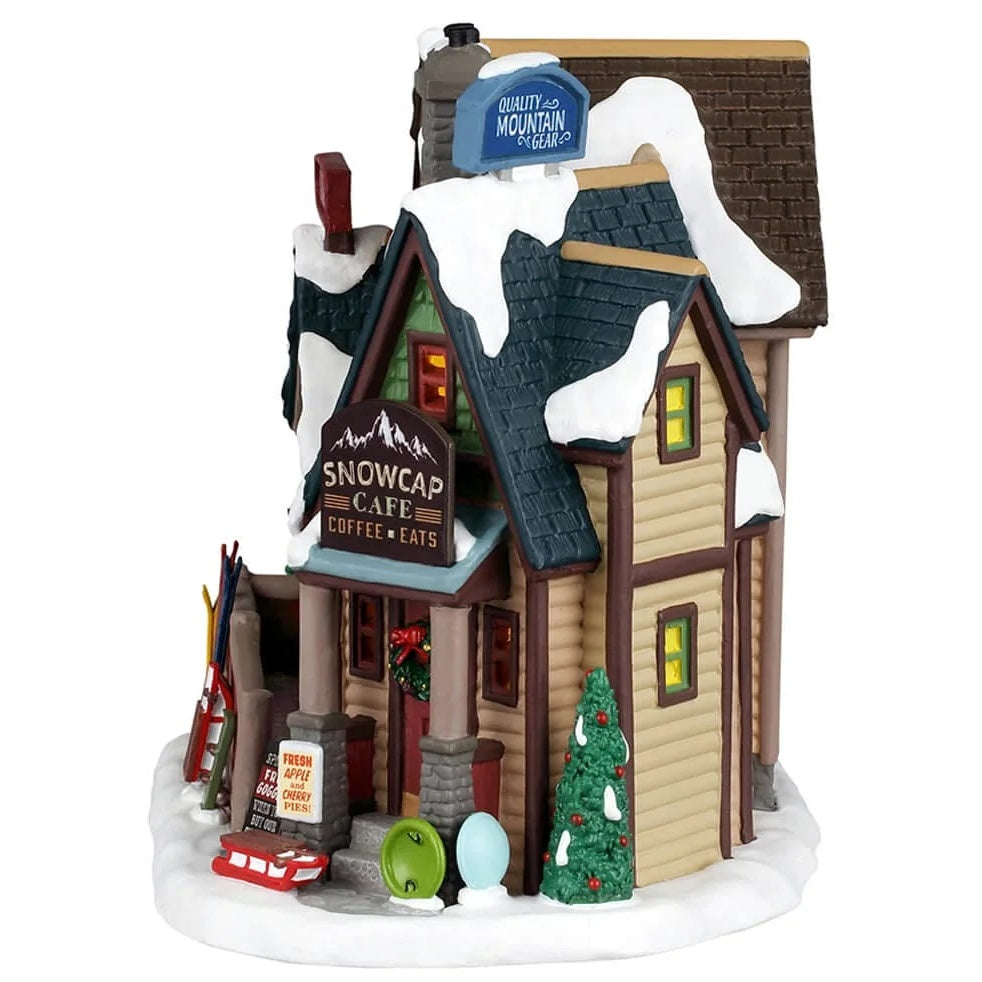 This image shows one of the sides of the building. There are signs on the building that read "Quality Mountain Gear" and "Snowcap Cafe Coffee & Eats." The roof and ground are covered in snow. Near the front door, there are some tubes for sledding.