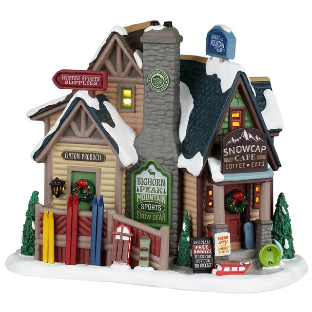 This image shows what this Lemax building looks like on a white background. It looks like it was a large log cabin with a stone chimney that has been turned into a bunch of different shops. Outside around the porch, there are skis, sleds and snowboards leaning, waiting for people to come out and use them. There are signs that read "Winter Sports Supplies," "Custom Products," "Bighorn Peak Mountain Sports Snow Gear," "Special! Free goggles when you buy our ski package" and "Fresh apple and cherry pies!"