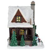 This image shows what the side of this cabin looks like. The roof and ground are both covered in snow. There's a squirrel standing near one of the trees.