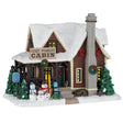 This image shows what this Lemax cabin looks like on a white background. It was designed to look like a log cabin covered in snow. Near the front porch, there are a bunch of trees covered in snow and three snow people decorated with hats and scarves. The chimney is made of stone and there's a decorated wreath hanging on it. On the house, on either side of the chimney, there's a red bow.