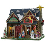 This image shows what this Lemax building looks like on a white background. It was designed to look like an old-timey cottage turned into a Christmas decoration shop. There are decorative snowflakes on the roof, cardboard cutout decorations in front of the shop and people inside working.