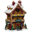 This image shows what this Lemax building looks like on a white background. It was designed to look like a tall log cabin-themed hotel. There's snow on the roof and the sidewalk. There are three gold stars above the front door. The front doors are decorated with red bows. There are trees on either side of the front door and skis leaning against the wall.