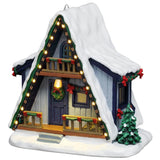 This image shows what this Lemax Vail Village table accent looks like on a white background. This piece was designed to look like an A-frame winter cabin. It's a blue wooden house with lights lining the roofline. The balcony, porch and the sides of the house are all decorated with green garland and red bows. There's a wreath hanging on the door and there's snow all over the house, trees and ground.