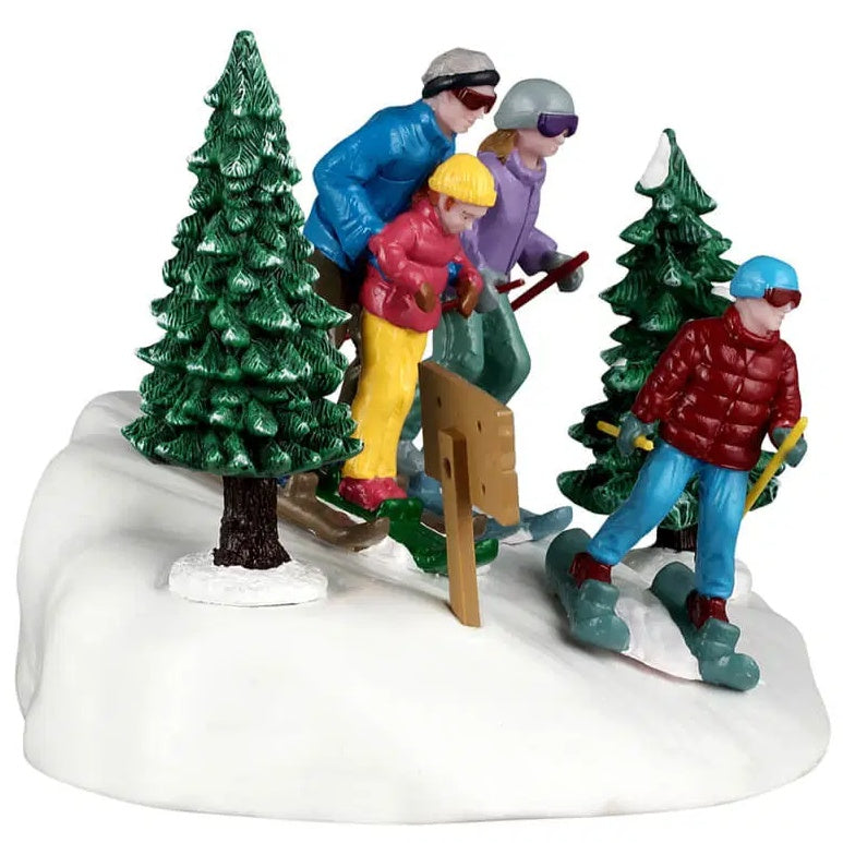 This image shows the side of this table accent. The dad is wearing a gray and black hat, brown goggles, a blue coat, black pants and brown skis. The mom is wearing a gray hat, purple goggles, a purple coat, green pants and blue skis. The dad is holding a young girl and helping her ski. She is wearing a yellow hat, a red coat, yellow pants, red boots and green skis. The young boy is wearing a blue hat, red goggles, a red coat, blue pants, red shoes and teal colored skis.