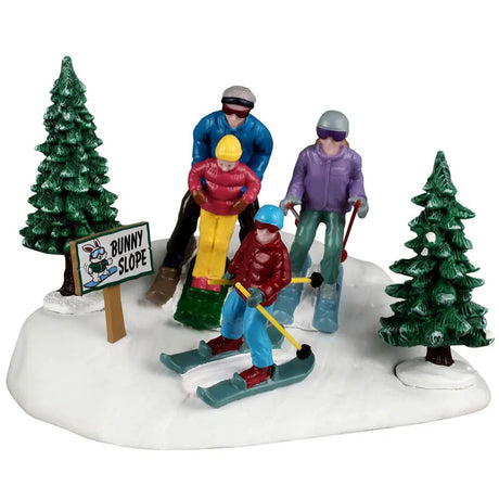 This image shows what this Lemax Vail Village table accent looks like on a white background. It's designed to look like a family skiing through snow in a forest. There's a sign that reads "Bunny Slope."