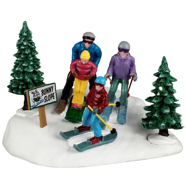 This image shows what this Lemax Vail Village table accent looks like on a white background. It's designed to look like a family skiing through snow in a forest. There's a sign that reads "Bunny Slope."