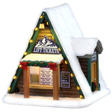 This image shows what this Lemax Vail Village table accent looks like on a white background. This piece was designed to look like an A-frame winter cabin. It's a log house with lights lining the roofline. There's a window where you can order tickets for the lift and a few menus with prices. There's also a sign that reads "Ask About Our All-Season Pass."