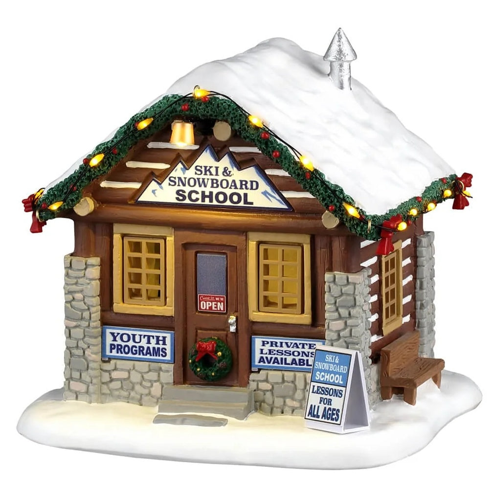 This image shows what this Lemax building looks like on a white background. It was designed to look like a ski and snowboard school.
 rental cabin. The roofline is decorated with lights and a decorated garland. There's an open sign on the door, as well as a decorated wreath. There are two signs underneath the front windows. One reads "Youth Programs." The other sign reads "Private lessons available." There's also a sign on the sidewalk that reads "Ski & Snowboard School: Lessons for All Ages."