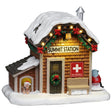 This image shows what this Lemax table accent looks like on a white background. It was designed to look like a small ski patrol station. The roof and the ground is covered in snow. There's a small dog house on the left side and a red snowmobile on the right side. There's a dog in the dog house wearing a Santa hat. There's a wreath on the front door and a garland decorated with red bows along the roofline. There's a bench next to the front door and a sign that reads "SKI PATROL."