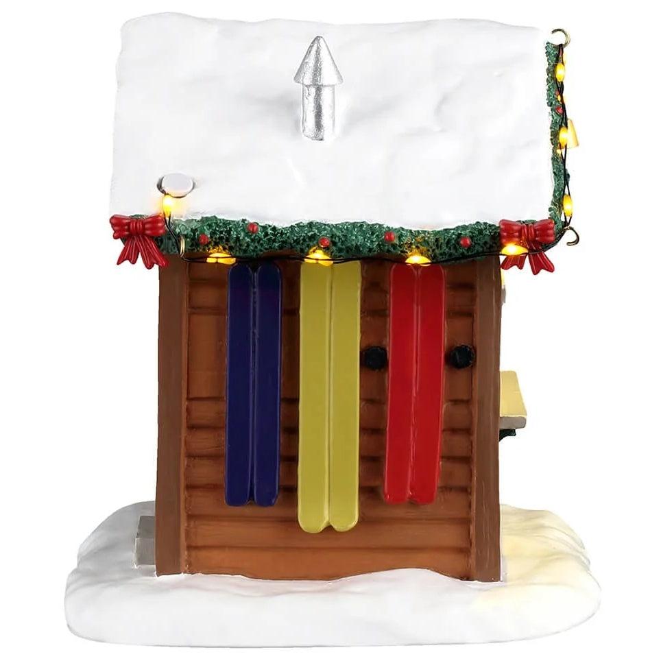 This image shows what the other side of this building looks like. The roof is covered in snow. There are three pairs of skis hanging on the wall: one set is dark blue, one set is a pale yellow and one set is red.