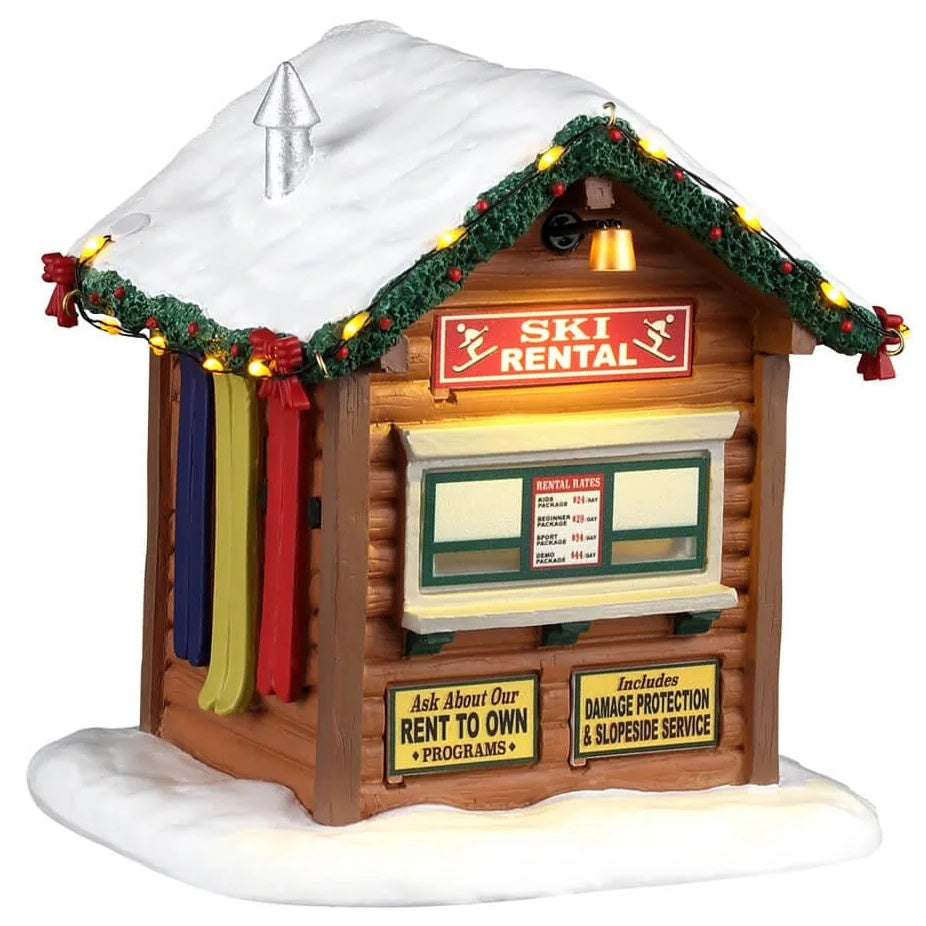 This image shows what this Lemax building looks like on a white background. It was designed to look like a ski rental cabin. There's a window with a counter. There's a price list in the window and there are two signs under the counter. One sign reads "Ask about our rent to own programs." The other sign reads "Includes damage protection and slopeside service." The roofline is decorated with lights and a decorated garland.