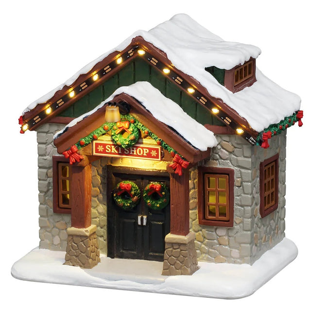 This image shows what this Lemax table accent looks like on a white background. It was designed to look like a small ski shop with stone sidings. The roof and the ground is covered in snow. There are two wreaths on the front doors and a garland decorated with red bows along the roofline.