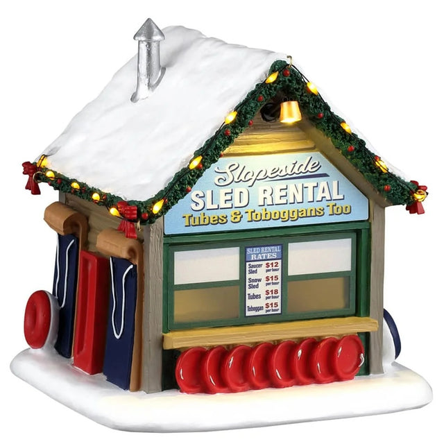 This image shows what this Lemax building looks like on a white background. It was designed to look like a small sled rental cabin. The front has a window with a counter and underneath the counter, there are a bunch of round sleds. There's a price list hanging in the window. Above the window there's a sign that reads "Slopeside Sled Rental: Tubes & Toboggans Too." There's snow on the roof.