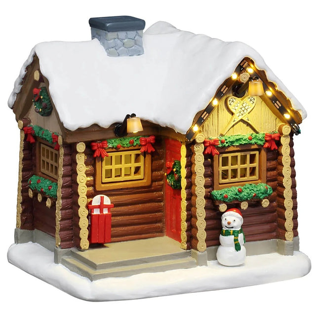 This Lemax Vail Vail Village table accent was designed to look like a small log cabin covered in snow. The windows are decorated with green garlands and red bows. There's a small snowman to the right of the front door. The snowman is wearing a red hat and a green scarf.