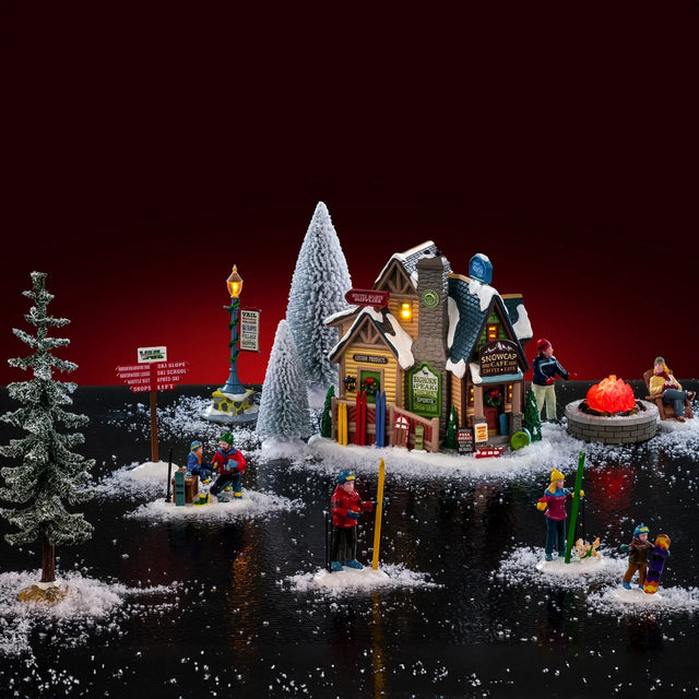 This is a lifestyle image showing this Lemax house set n on a fake background. This set was designed to look like a winter cabin where people are getting ready to start skiing and snowboarding. There's also a couple sitting next to a bonfire.