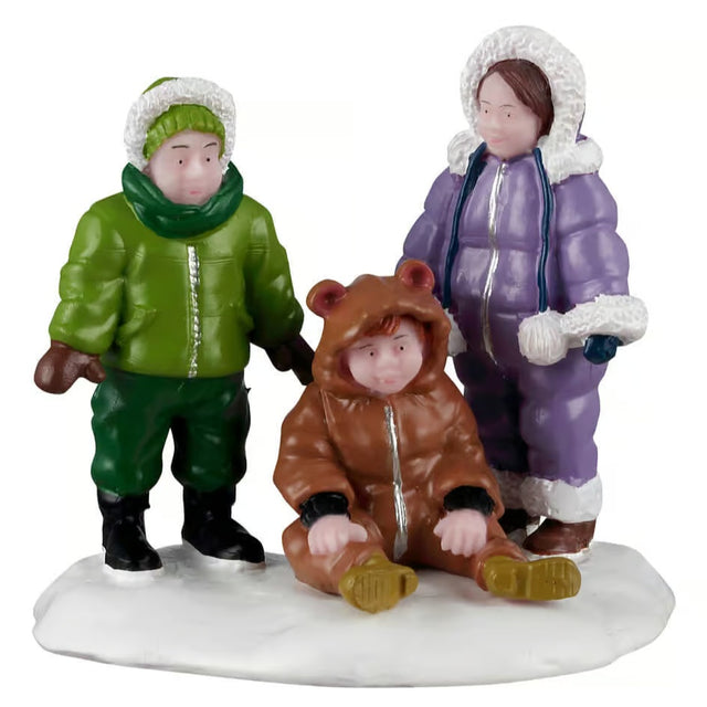 This image shows what this Lemax figurine looks like on a white background. It was designed to look like three kids wearing winter clothes in the snow. There's a little boy wearing a green hat, green scarf, green jacket, brown gloves, green pants and black boots. There's a little kid sitting on the ground wearing a onesie that looks like a bear. And there's a little girl wearing a purple and white snowsuit.