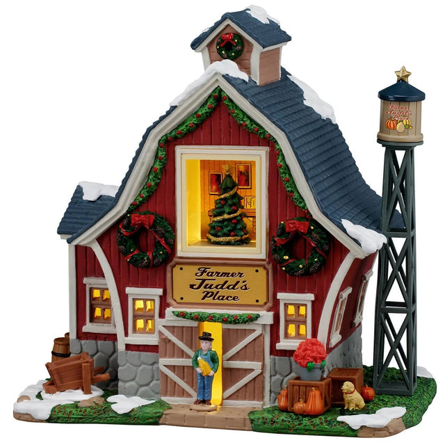 This image shows what this Lemax building looks like on a white background. It was designed to look like a red farmhouse that's been decorated for Christmas. There are wreaths and garlands and poinsettias all over. There's a large window in the center of the house and you can see a decorated Christmas tree. Farmer Judd is standing near the front door and there's a cute dog standing next to a few pumpkins.