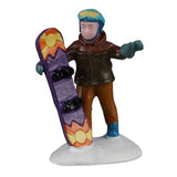 This image shows the little boy figurine standing in a pile of snow. He's holding a snowboard that is purple with a yellow and orange sun on it. He's wearing a blue hat, a brown jacket, brown pants, blue gloves and blue boots.