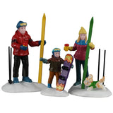 This image shows what these three Lemax Vail Village figurines look like on a white background. They were designed to look like a family about to enjoy time in the snow. The mom and dad have skis and the young boy has a snowboard. There's also a dog wearing a green sweater running around.