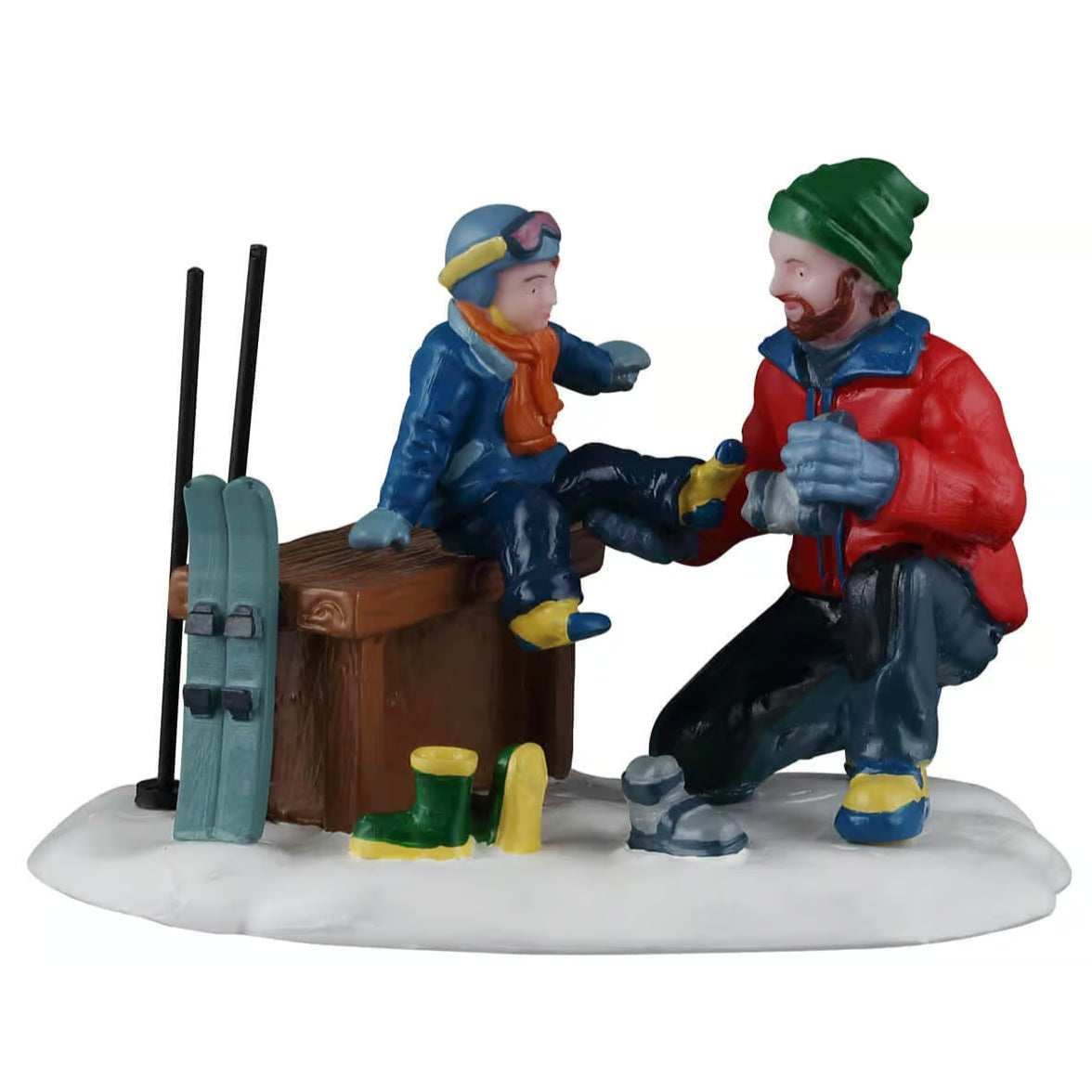 This image shows what this Lemax Vail Village figurine looks like on a white background. It was designed to look like a dad helping his young son put on his shoes to go skiing. The boy is sitting on a wooden bench. There are small skis leaning up against the bench.