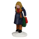 This image shows what the young girl figurine looks like on a white background. She has blonde hair and is wearing a blue and brown jacket, brown pants and black boots. She's holding a green bag and a red bag.