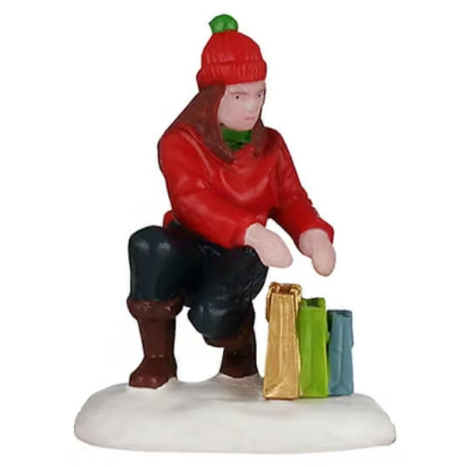 This image shows what the mom figurine looks like on a white background. She's wearing a red hat, a green scarf, a red sweater, blue pants and brown boots. She's leaning down checking her shopping bags. There's a gold bag, a lime green bag and a small blue bag.