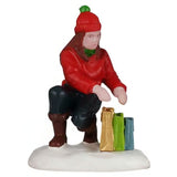 This image shows what the mom figurine looks like on a white background. She's wearing a red hat, a green scarf, a red sweater, blue pants and brown boots. She's leaning down checking her shopping bags. There's a gold bag, a lime green bag and a small blue bag.