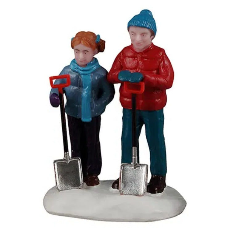 This image shows two kids standing in snow holding shovels. The young girl on the left has her hair in two buns and is wearing a blue puffy coat, a blue scarf, black pants and brown shoes. The young boy on the right is wearing a blue knit hat and a red puffy coat, blue jeans and black shoes.