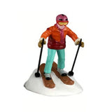 This image shows one of the figurines. It was designed to look like someone skiing downhill. They're wearing a red helmet with red goggles, an orange/red jacket, a red scarf, teal pants, yellow gloves, brown skis and black ski poles.