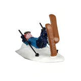This image shows one of the figurines. It was designed to look like a person who fell backwards while skiing. They're laying on their back in the snow with their skis in the air. They're still holding onto their ski poles. They're wearing a red hat, goggles, a blue jacket and blue pants.